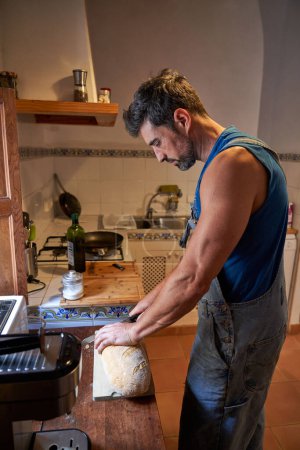 Téléchargez les photos : Vue latérale de l'homme barbu concentré en salopette de denim debout près du comptoir et couper le pain tout en préparant le petit déjeuner dans la cuisine le matin - en image libre de droit