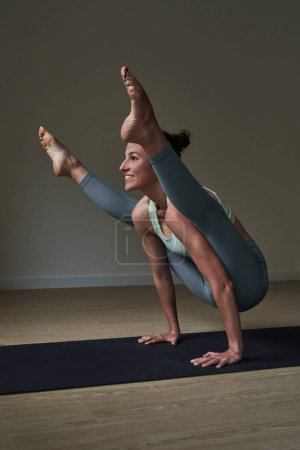Téléchargez les photos : Corps complet d'athlète féminine souriante en vêtements de sport prenant la pose Tittibhasana sur un tapis noir tout en pratiquant le yoga dans un club moderne - en image libre de droit