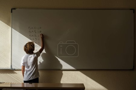 Foto de Vista posterior de un colegial anónimo escribiendo tabla de multiplicación en pizarra blanca en el aula durante la lección de matemáticas - Imagen libre de derechos