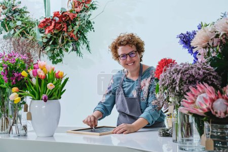 Téléchargez les photos : Fleuriste professionnelle souriant et regardant la caméra tout en se tenant à table avec des fleurs colorées en fleurs et en écrivant à bord avec de la craie pendant la journée de travail dans un magasin de fleurs moderne - en image libre de droit