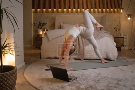 Téléchargez les photos : Femme en tenue de sport faisant de la gymnastique avec ordinateur portable dans une chambre moderne aux couleurs pastel - en image libre de droit