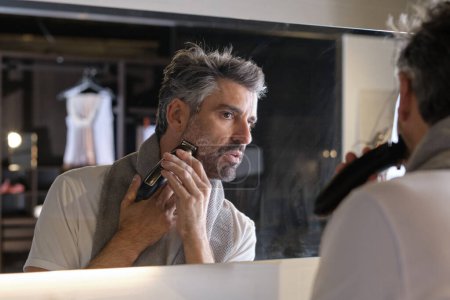 Photo for Focused man with gray beard doing morning routine and shaving using professional trimmer while looking at mirror in modern bathroom at home - Royalty Free Image