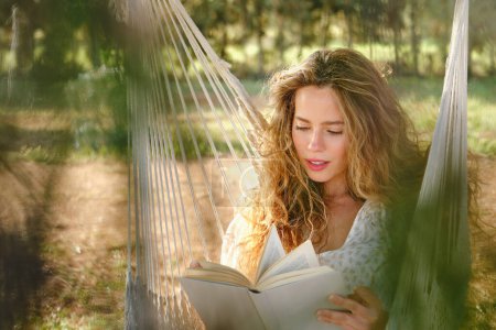 Photo for Young woman with curly hair sitting on hammock and reading interesting book on sunny weekend day in garden - Royalty Free Image