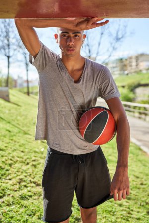Foto de Ajuste jugador de baloncesto étnico en ropa deportiva de pie con pelota cerca de la construcción en el parque verde y mirando a la cámara - Imagen libre de derechos