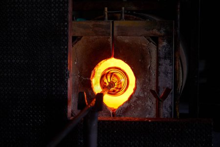 Foto de Objeto de vidrio en la punta de la tubería colocada en horno de combustión en instalaciones oscuras en la fábrica de soplado de vidrio - Imagen libre de derechos