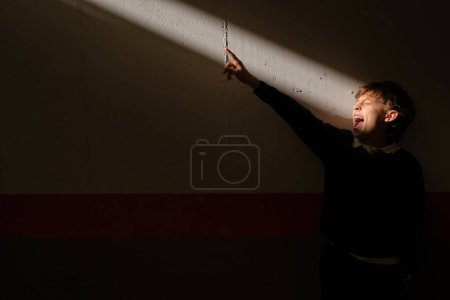 Téléchargez les photos : Mignon preteen garçon debout dans sombre chambre à mur attrapant rayon de soleil et criant et pointant doigt à fenêtre - en image libre de droit