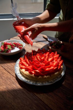 Photo for Anonymous cook standing at wooden table and garnishing pie cake with tomato sauce while decorating crust with strawberry fruit slices placed in daylight - Royalty Free Image