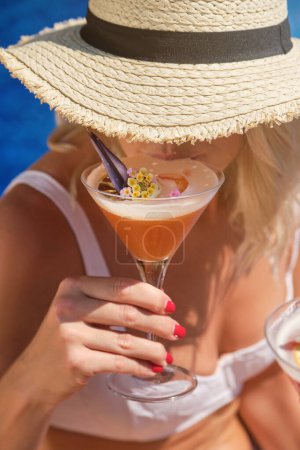 Téléchargez les photos : Femme méconnaissable en chapeau de paille et maillot de bain blanc boire cocktail rafraîchissant tout en se reposant pendant la journée d'été - en image libre de droit