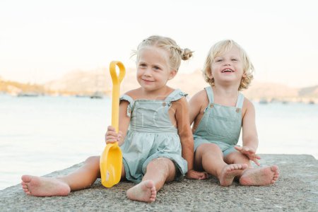 Photo for Full body of cheerful little children in casual clothes sitting on stone border with toy shovel at beach on sunny day - Royalty Free Image