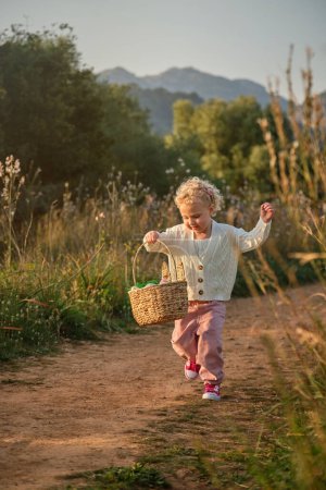 Téléchargez les photos : Petite fille adorable pleine longueur en pull doux portant un panier en osier et se promenant sur un chemin de terre dans une campagne paisible - en image libre de droit