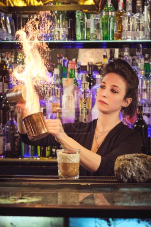 Photo for Concentrated skilled young female bartender with dark hair adding salt into metal cup while mixing flaming cocktail in modern bar - Royalty Free Image