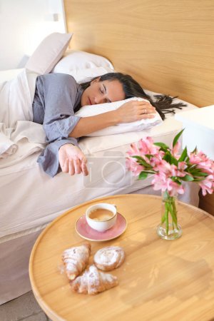 Téléchargez les photos : Femme endormie en pyjama couchée sur un lit doux près d'une table en bois avec croissant et bouquet de fleurs fraîches dans la chambre à coucher à la maison - en image libre de droit