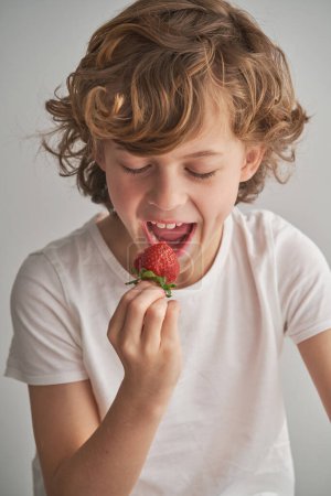 Foto de Alegre niño despreocupado con los ojos cerrados mordiendo bayas de vitamina dulce sobre fondo gris - Imagen libre de derechos