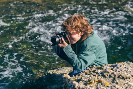 Foto de Desde arriba vista lateral de niño alegre tomando fotos de la naturaleza con cámara fotográfica en la orilla rocosa cerca del río que fluye - Imagen libre de derechos