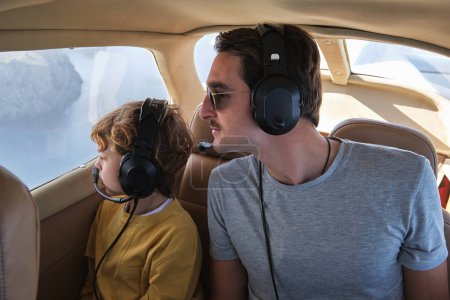 Téléchargez les photos : Jeune homme et fils en vêtements décontractés et écouteurs volant dans l'avion et regardant par la fenêtre avec curiosité pendant le voyage - en image libre de droit