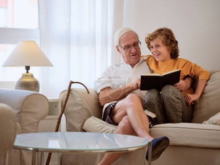 Téléchargez les photos : Délicieux homme âgé avec petit-fils positif lisant livre intéressant ensemble tout en étant assis sur le canapé dans le salon de lumière à la maison - en image libre de droit
