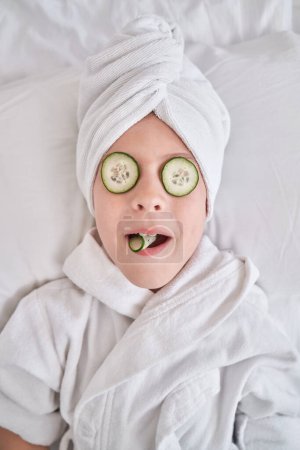 Foto de Chico divertido con máscara de pepino en los ojos con turbante de toalla y albornoz descansando en la cama blanca en casa durante la rutina de belleza de cuidado de la piel - Imagen libre de derechos