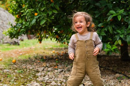 Téléchargez les photos : Contenu enfant en salopette riant sur le terrain de galets et regardant loin contre un arbre luxuriant avec des oranges dans la campagne - en image libre de droit