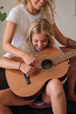 Téléchargez les photos : Photo verticale d'une fille blonde jouant de la guitare tout en souriant avec une femme assise avec elle sur un canapé dans une pièce - en image libre de droit