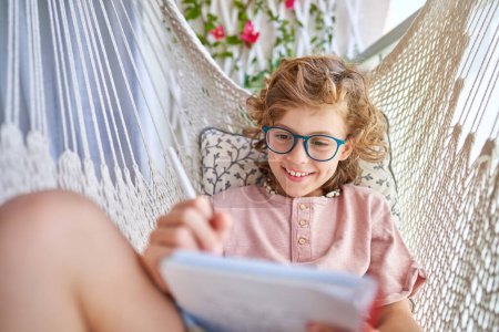 Photo for Smiling child in eyeglasses resting in knitted hammock with cushion and writing in diary with pencil on balcony at home - Royalty Free Image