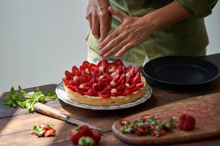Téléchargez les photos : Cuire méconnaissable en tablier vert découpant délicieux gâteau aux fraises sur une assiette avec couteau - en image libre de droit