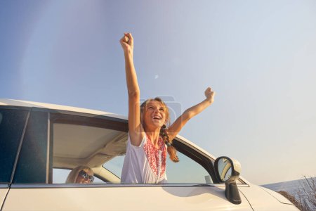 Photo for Blonde girl sticking her body out of the window of a car with the arms raised in gesture of happiness with her mother sitting inside the car - Royalty Free Image