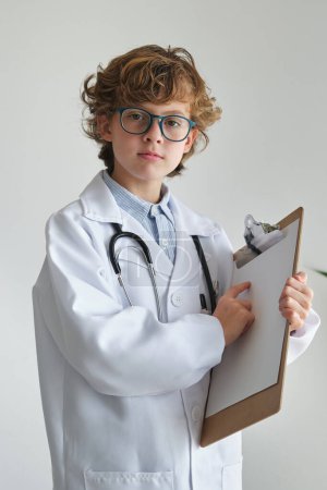 Photo for Smart child in eyewear and medical uniform pointing at empty paper on clipboard while looking at camera on white background - Royalty Free Image