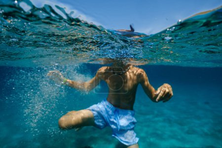 Foto de Vista submarina del niño anónimo sin camisa en pantalones cortos sumergiéndose en agua de mar ondulante azul durante las vacaciones de verano - Imagen libre de derechos