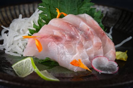 Téléchargez les photos : Gros plan de délicieux sashimi de bar cru servi sur une assiette noire avec des fleurs de laitue verte et du radis sur fond flou - en image libre de droit