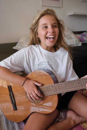 Foto de Chica positiva emocionada con sonrisa dentada y en camiseta blanca tocando la guitarra acústica mientras está sentado en el suelo en la sala de estar - Imagen libre de derechos