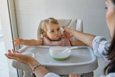 Foto de Mujer de la cosecha limpiando la cara sucia de niña curiosa en babero sentado en la silla alta con plato de comida en la cocina casera - Imagen libre de derechos