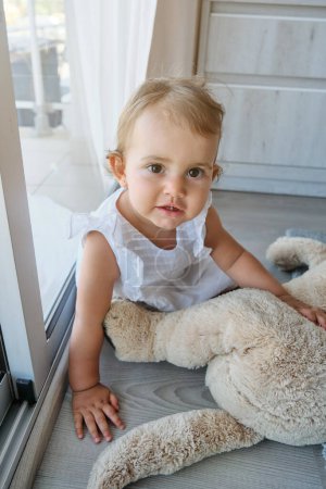 Foto de Alto ángulo de bebé curioso en vestido blanco jugando con peluche y mirando a la cámara con interés mientras está sentado en el suelo cerca de la ventana en la sala de estar en casa - Imagen libre de derechos
