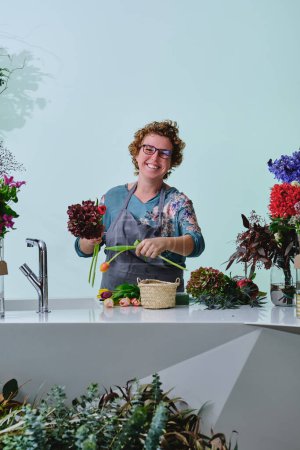 Téléchargez les photos : Joyeux fleuriste féminin d'âge moyen avec des cheveux bouclés courts dans des lunettes arrangeant bouquet de diverses fleurs et regardant la caméra dans la boutique de fleurs - en image libre de droit