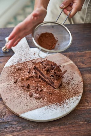Photo for High angle of crop anonymous person sprinkling cocoa powder with strainer over delicious homemade chocolate cake on board at table - Royalty Free Image