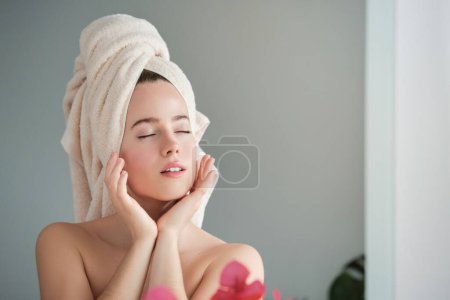 Téléchargez les photos : Charmante jeune femme aux cheveux enveloppés dans une serviette blanche rêvant après avoir fait la routine beauté du matin dans le salon à la maison - en image libre de droit