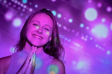 Téléchargez les photos : Joyeux jeune femme serrant les mains et souriant joyeusement tout en faisant vœu lors de la célébration des vacances sous la projection lumineuse de taches de lumière - en image libre de droit