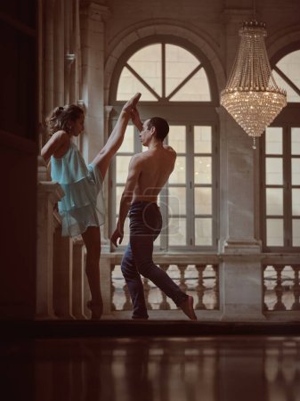 Foto de Vista lateral del hombre descalzo sin camisa ayudando a la suave bailarina haciendo de pie dividida en salón clásico - Imagen libre de derechos