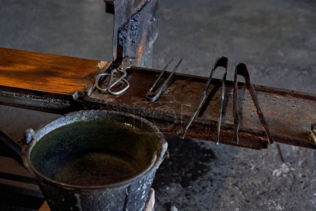 Téléchargez les photos : Divers outils de soufflage du verre sur un banc en bois près d'un seau rempli d'eau dans une usine de fabrication de verre - en image libre de droit