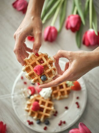 Photo for From above crop anonymous person holding tasty homemade waffle with berries while standing at table with tulips in light kitchen - Royalty Free Image