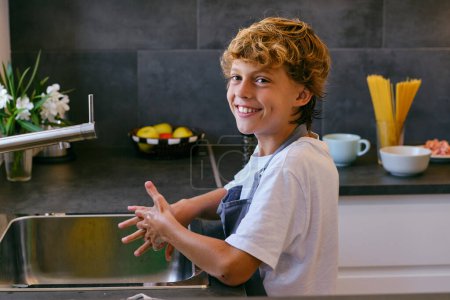 Téléchargez les photos : Vue latérale de l'enfant joyeux avec les cheveux bruns regardant la caméra tout en se lavant les mains au-dessus de l'évier dans la cuisine de la maison - en image libre de droit