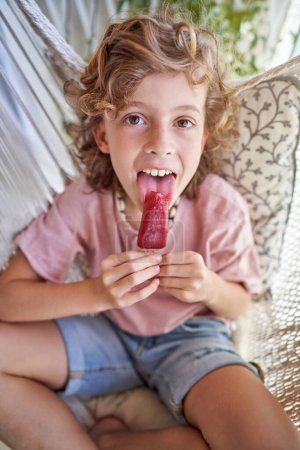 Hübscher Junge mit kurzen lockigen Haaren in Jeans und rosa T-Shirt leckt süßes gefrorenes Eis und schaut in die Kamera, während er im Sommer in der Hängematte sitzt