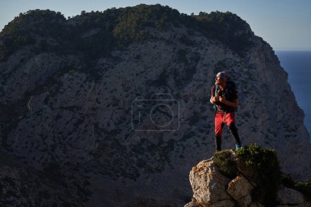 Photo for Full body of bearded male traveler with backpack standing on stony top in highlands and observing majestic landscape under cloudless blue sky - Royalty Free Image