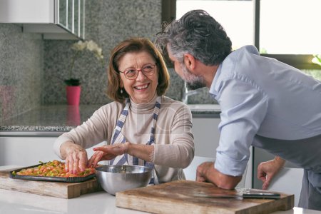 Foto de Mujer madura positiva en gafas y hombre sin afeitar cocinar pizza juntos y divertirse mientras se miran el uno al otro en el día - Imagen libre de derechos