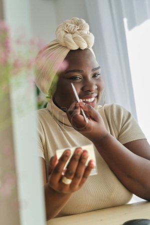 Téléchargez les photos : Jeune femme noire dans le turban africain traditionnel appliquant eye-liner noir et souriant tout en regardant dans le miroir - en image libre de droit