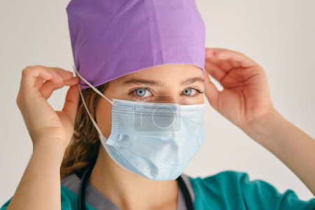 Téléchargez les photos : Médecin professionnel en peignoir médical touchant la tête tout en mettant un masque de protection et en regardant la caméra sur fond beige - en image libre de droit