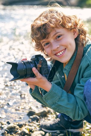 Photo for Positive boy taking picture with photo camera looking at camera while sitting on haunches on coast near flowing water on blurred background - Royalty Free Image
