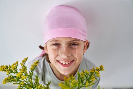Photo for Kid in pink headband looking at camera with tooth smile near Solidago against white background - Royalty Free Image