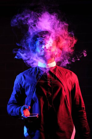 Photo for Black male with smartphone in hand vaping against black background in red and blue neon lights with fume - Royalty Free Image