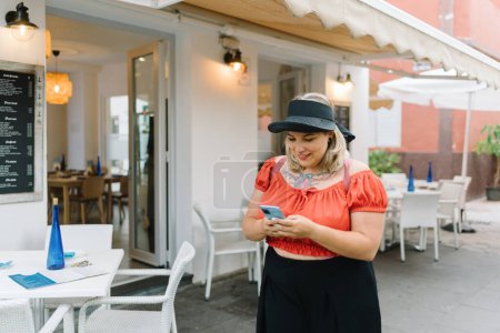 Photo for Positive young plus size female in stylish outfit and hat smiling browsing smartphone while standing in street cafe with white wall and furniture - Royalty Free Image