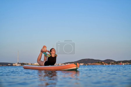 Téléchargez les photos : Corps complet de jeune femme flexible en maillot de bain pratiquant le yoga à Dhanurasana sur la planche SUP entourée d'une mer ondulante contre un paysage pittoresque - en image libre de droit